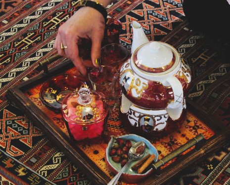 Hands serving traditional Persian tea in decorative glass cups, cinnamon and other spices on small plates, Persian carpet in background
